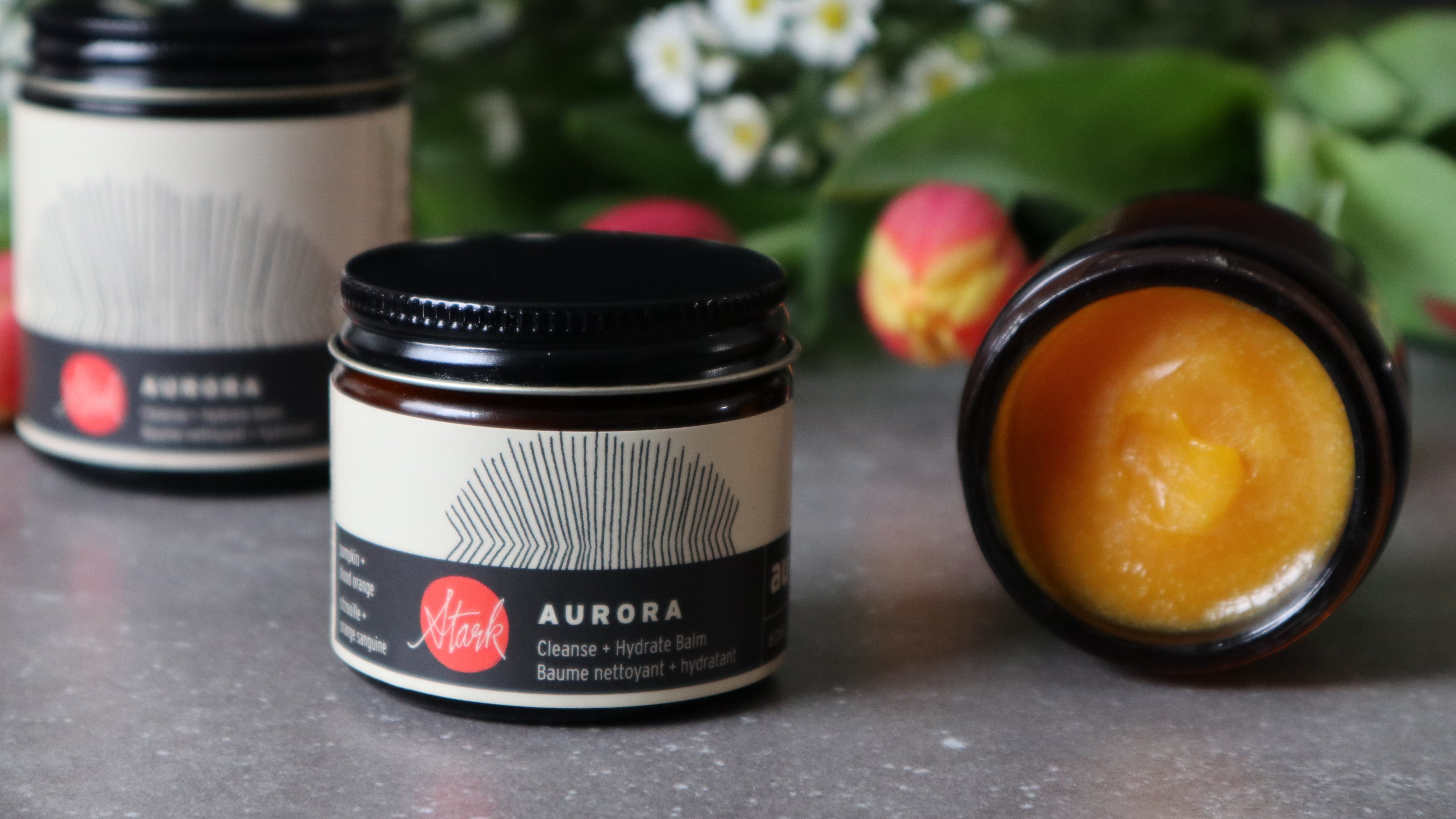 Jars of a product with 'Aurora' branding on a surface with flowers in the background